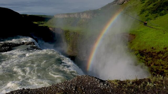 Experience the breathtaking beauty of Gullfoss waterfall in Iceland as a vibrant rainbow arches over rushing waters. The lush greenery contrasts with the powerful cascade, creating a mesmerizing view.