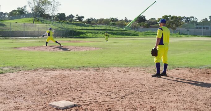 Following windup, teammates in yellow uniforms pitching from mound and tracking ball on park field