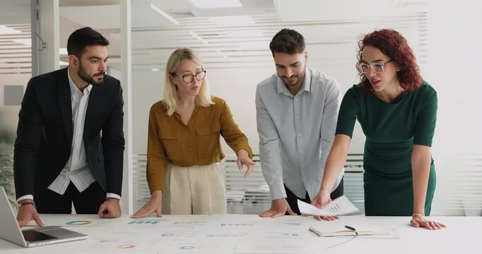 Four multinational teammates reviewing financial data, analyzing charts displayed on sprinted documents spread out on table, discussing key metrics, collaborating on strategy. Teamwork, strategizing
