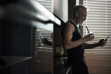 Middle aged Caucasian man standing in kitchen holding smartphone and drinking smoothie, smiling while looking at screen, sunlight streaming through window blinds in background