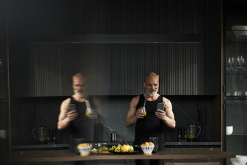 Middle aged Caucasian man standing in modern kitchen holding smartphone and glass, smiling while reading message, muscular build visible, fresh fruits and vegetables on counter