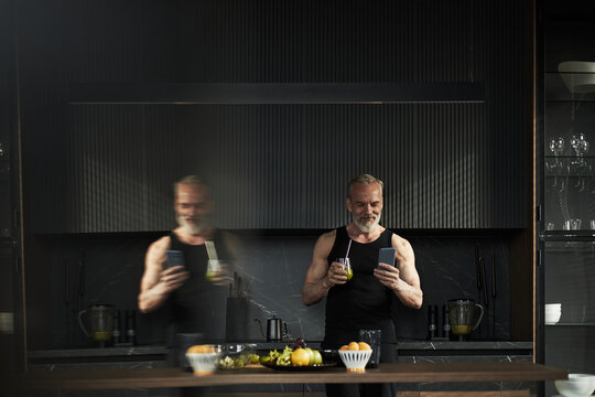 Middle aged Caucasian man standing in modern kitchen holding smartphone and glass, smiling while reading message, muscular build visible, fresh fruits and vegetables on counter - Powered by Adobe