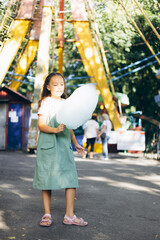 Obraz premium Young girl enjoying cotton candy at an amusement park on a sunny day, surrounded by vibrant attractions.