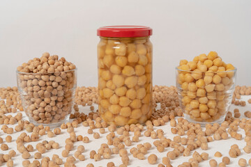 Raw chickpeas. Preserved chickpeas. Cooked chickpeas. All in clear glass containers. On a white table. Loose chickpeas around them.
