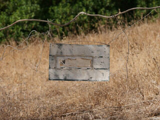 a weathered, rectangular sign with no content hanging on a wire fence, with blurred areas of dry grass and green vegetation visible in the background