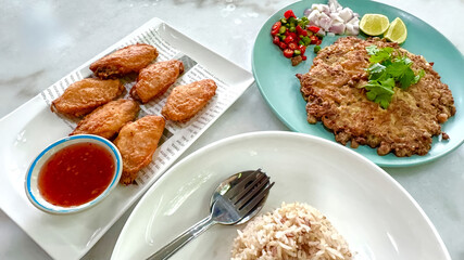 A meal consisting of fried chicken wings served with sweet chili sauce, a Thai style omelet minced pork and salted fish with herbs and lime on the side, and a plate of rice served.