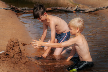 Fototapeta premium Shirtless boys play in muddy water, building a long mud structure on a sandy riverbank in bright summer sunshine. A playful and creative outdoor scene.