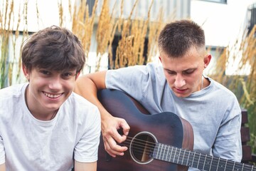 Fototapeta premium Two young people relaxing outdoors with a guitar. A young man is playing the guitar while a teenage boy listens cheerfully. A warm and joyful moment of friendship and music.