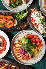 Assorted gourmet dishes on plates, featuring grilled chicken salad, smoked salmon fritters, tomato cheese salad, and a creamy dessert with mint garnish