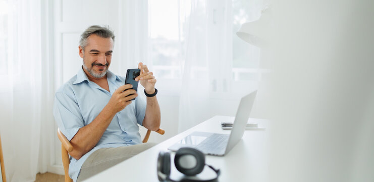Confident mature male professional smiling and multitasking with a laptop and mobile phone in a workspace.