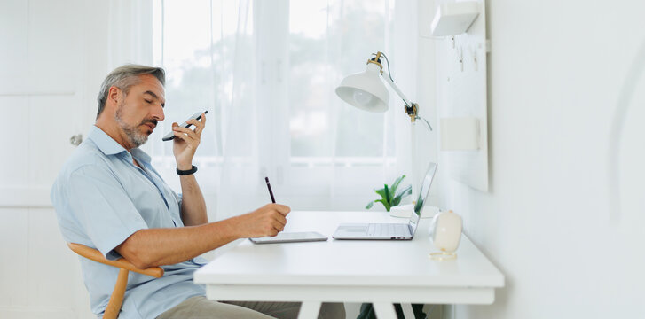 Middle-aged man writing down notes on digital tablet at a white desk while multitasking with his smartphone and laptop.
