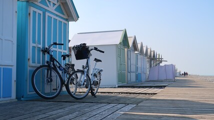 Cyclotourisme dans la baie de Somme, en Picardie / Hauts de France, deux vélos garés côte à côte devant une cabine de plage, sur les planches de la ville de Cayeux sur Mer (France)
