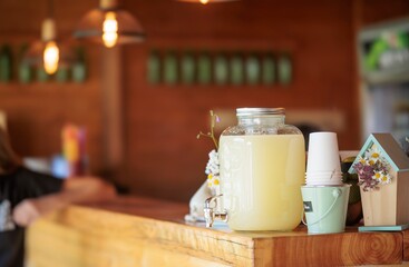 At a local cafe, a large jar of lemonade sits on a wooden counter, ready to be dispensed into stacked cups. Flowers and a decorative house add a touch of country charm