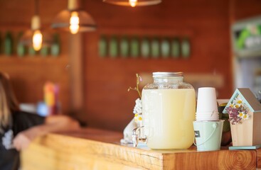 Lemonade in a large jar sits on a wooden counter at a cheerful lemonade stand during a summer fair. A stack of cups and a decorative birdhouse complete the summery display