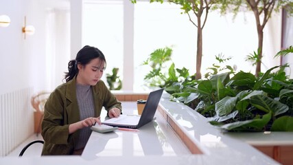 Asian adult woman freelancer calculates business finances using laptop calculator at bright modern cafe workspace