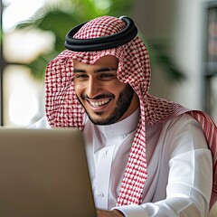 An Arab man in traditional dress smiles while sitting at a laptop.