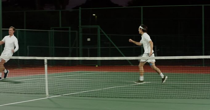 Diverse male tennis partners volleying net shots under lights after lobbed yellow ball, celebrating