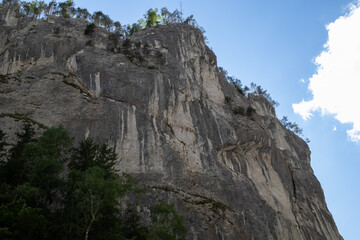 limestone cliff face, rugged texture, forest at base, clear blue sky, white clouds, natural outdoor rock formation, dramatic landscape, vertical perspective