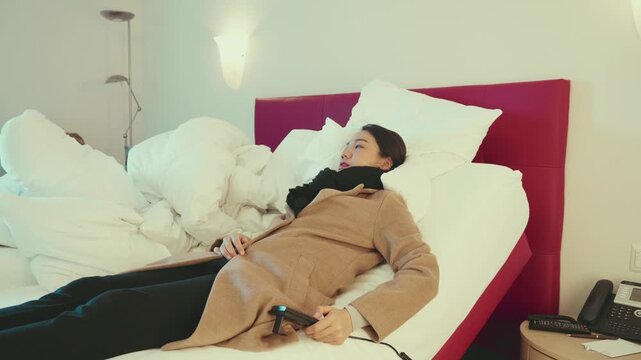 A woman lounges comfortably on a hotel bed, surrounded by soft white pillows and blankets. She appears relaxed, holding a remote control, enjoying her time in a stylishly decorated room with a red acc