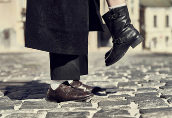 A playful moment in urban fashion, with a pair of legs in brown shoes standing on a cobblestone street while another person's leg in a black boot is lifted in the air. This image captures a stylish, l