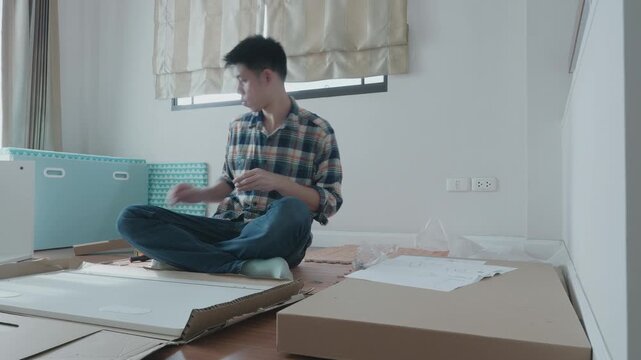 A young man assembles furniture in a well-lit room, surrounded by packaging and tools. The scene captures the focus and determination as he puts together the pieces, showcasing a moment of DIY creativ