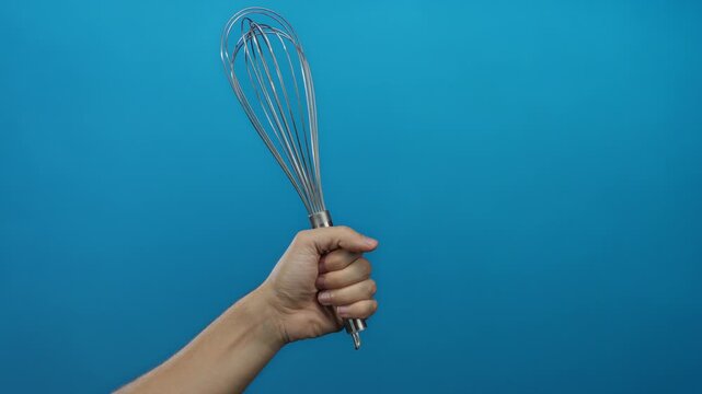 Caucasian man holding metal whisk isolated against blue wall background showcasing kitchen tool and cooking concept with simplicity and focus.