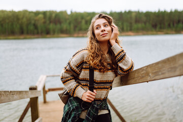 Portrait of a young female traveler on a wooden walking path overlooking a lake. Cute woman with a backpack enjoying nature and walking near the forest. Adventure, nature concept.