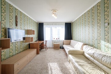 bright living room with patterned wallpaper, a light carpet, a beige sofa, a wooden desk, and a TV.  The room has a window with dark curtains