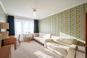 brightly lit living room features a beige sofa, a wooden desk near a window, and striped wallpaper. The room is carpeted and appears cozy