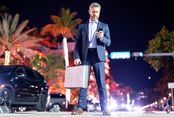 Fototapeta premium Businessman with briefcase and phone walking at city. Rich man in suit in night. Businessman near luxury car. Businessman walking near night city. Business style. Rich businessman in luxury setting.