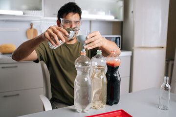 Home gardening enthusiast wearing safety glasses carefully pouring liquid from glass bottle through funnel into plastic bottle, creating plant fertilizer solution in kitchen setting.