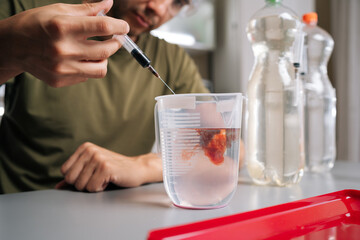 Close-up cropped shot of chemist in safety glasses adding red liquid fertilizer to water in beaker using syringe, preparing solution for plant growth and nutrition. Concept of home gardening.