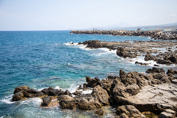 Rocky shoreline, waves splashing on jagged stones and concrete block breakwater in Rethymno, Crete, Greece.