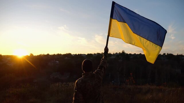 Male military in uniform lifting blue-yellow banner in honor of the victory against russian aggression at sunset. Young soldier of ukrainian army raised a waving flag of Ukraine at countryside