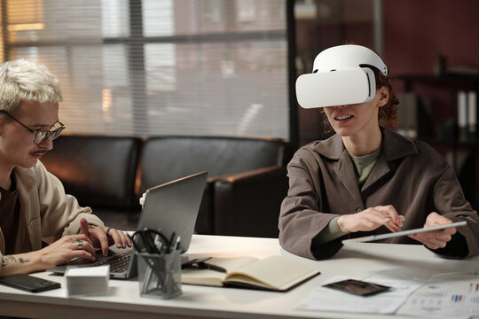Young female manager using digital tablet and VR headset while her male colleague typing on laptop in modern office