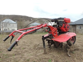 red hand petrol plow standing on farm land with dry grass against background of village houses, mechanization of private farming with special equipment for soil cultivation