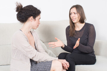 Two women sitting on a couch having a heated argument, one gesturing emotionally while the other listens with tension