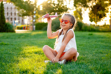 a little child girl on a green lawn flying a plane, a laughing child in red glasses on the grass in summer, summer vacation