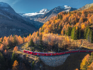 Aerial view of red Bernina Express train passing through colorful autumn forest in the Swiss Alps with high mountains, glacier, curved railway on a scenic sunny day. Top drone view of train in fall