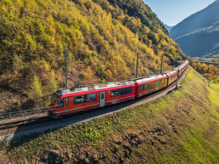 Aerial view of modern red train in alpine mountains, green meadows, orange trees at sunset in autumn. Bernina Express, Switzerland. Top view of train in Alps, winding railroad in fall. Glacier Express