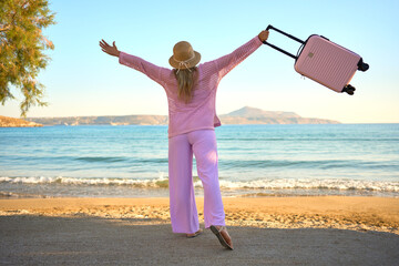 Happy woman with arms raised holding suitcase on sandy beach near sea. Excited tourist celebrating summer vacation travel.