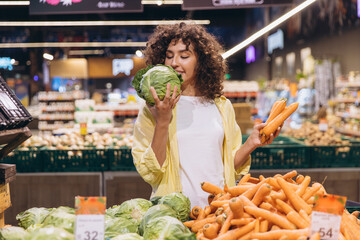 Woman smelling cabbage and holding carrots while shopping in supermarket