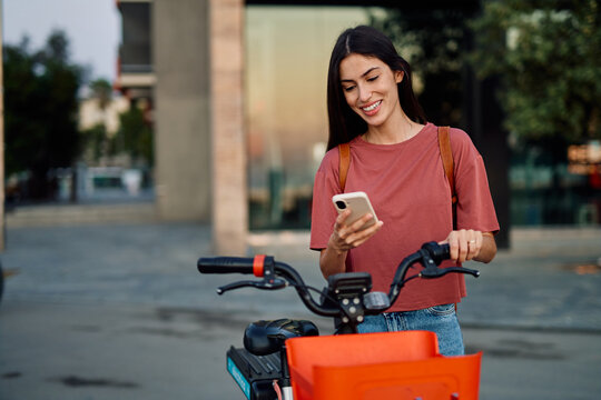Smiling happy beautiful woman using mobile phone to unlock electric bicycle in city