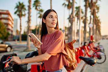 Smiling happy beautiful woman using mobile phone to unlock electric bicycle in city © Lumos sp