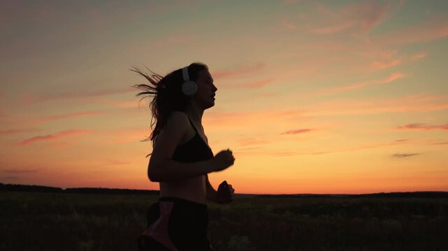 Silhouette, beautiful girl in musical headphones, is engaged in fitness, runs in evening. Training run. Free young woman runs in summer in park at sunset, listens to music on headphones. Teen runner - Powered by Adobe