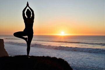 silhouette of a woman practicing a yoga tree pose on a cliff overlooking the ocean at sunset, representing peace, balance, wellness, and mindful meditation in nature
