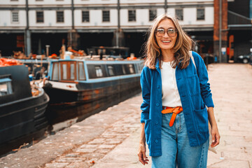 Young woman in blue cardigan and glasses smiling by canal with boats, enjoying a sunny day in urban...