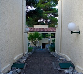 A narrow paved walkway between cream-colored buildings leads to a small courtyard with trees and globe-shaped lamps. The scene is framed by tall pine trees and Mediterranean architecture.
