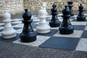 Large outdoor chess pieces in black and white stand on a checkered board against a stone wall. The...