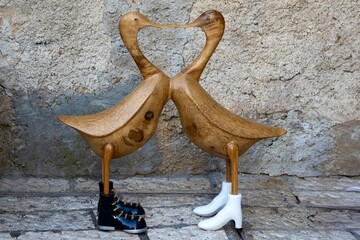 Two wooden duck figurines stand side by side, touching beaks in the shape of a heart. One wears white boots, the other black, creating a charming contrast.